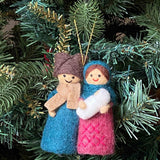 A Nativity Scene felt ornament hangs on a lush, green Christmas tree, evoking peace. This handmade felt ornament features a 3D design of the Holy Family in colorful felt. The man wears a brown hat, tan scarf, and blue robe, while the woman wears a blue shawl and pink robe with a criss-cross pattern, holding a small white figure. A gold cotton string is attached for hanging.