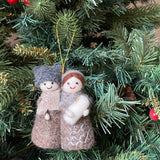 A handcrafted felt Nativity Scene ornament is suspended from a lush green Christmas tree, adorned with red berry decorations. This 3D felt ornament depicts Mary in a brown headscarf and gown, Joseph in a gray hat and robe, and baby Jesus wrapped in white, all beneath a shining star. A golden string suspends this unique Christmas ornament, adding faith and warmth to the holiday decor.