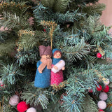 A handcrafted felt Nativity Scene ornament hangs on a Christmas tree adorned with green needles, small red berries, and gray, red, and white wool felt balls. This unique Christmas ornament features a 3D design of the Holy Family: Joseph in a brown hat and blue robe, Mary in a pink dress holding baby Jesus, and a shining star above, suspended by a gold string.