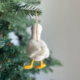 A handmade felt white duck ornament hangs from a pine tree branch in a festive Christmas tree setting, evoking a warm and joyful tone. This unique Christmas ornament features a fluffy, textured felt body and neck, complemented by bright yellow webbed feet. A jute string is attached for easy display.