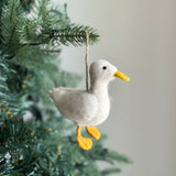 A charming white felt duck ornament rests among the lush green branches of a Christmas tree. This handmade wool felt ornament features a soft white body with a bright yellow beak and matching feet. A jute string allows the felt duck ornament to hang gracefully among the twinkling lights, adding a touch of handcrafted elegance.