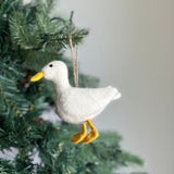 A felt white duck ornament hangs on a green Christmas tree, set against a soft, neutral background, evoking a cozy, festive mood. This handmade felt ornament features a textured white body, a small black eye, a yellow beak, and yellow webbed feet. A jute string is attached to the top for hanging.