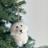 A white Maltese dog felt ornament hangs from a lush green Christmas tree partially adorned with small electric lights, creating a festive ambiance. This handcrafted felt Christmas ornament features a fluffy white texture and a cheerful expression, highlighted by its dark, beady eyes and a small, stitched mouth. A jute string is attached for easy hanging.