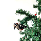 A felt woodpecker ornament hangs from a branch of a lush green Christmas tree, adding rustic charm to the festive setting. This handcrafted felt ornament is detailed in white, black, and red felt, showcasing realistic plumage and a distinctive beak. It is suspended by a jute string.