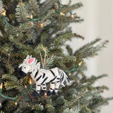 A charming needle-felted zebra ornament hangs on a green Christmas tree adorned with soft, warm lights. This delightful wool felt zebra ornament showcases classic black and white stripes, a playful fluffy mane, and a jute string for hanging, adding a festive touch to holiday decor.