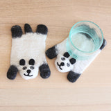 Two handcrafted felt panda coasters rest on a light wooden surface; one flat and one beneath a clear glass. Each charming wool felt panda coaster is primarily white with black accents on its ears, paws, and facial features, including a small smiling mouth and simple eyes. Crafted from soft 100% sheep wool, their handmade details create a warm, inviting charm, highlighting their ethical production by Nepalese female artisans.