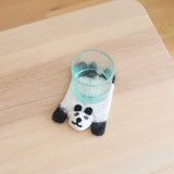 A light blue glass rests on a charming white felt panda coaster, part of a set, displayed on a light wood surface. This handcrafted wool felt panda coaster features a soft white body with black ears, legs, and a cute facial design. These ethically handmade felt panda coasters bring a playful touch to any home decor.