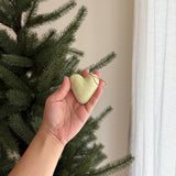 A single light green wool felt heart ornament is gently held in a female hand near a festive Christmas tree. This plump, needle-felted pastel heart ornament features a soft, uniform texture and a rustic jute string for hanging, illuminated by soft natural light.