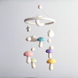 A Felt Pastel Mushroom Baby Mobile hangs against a pale gray backdrop, creating a whimsical and dreamy scene. This charming wool felt baby mobile features pastel mushrooms in pink, light green, blue, purple, and yellow, each with white polka dots. Fluffy white wool felt stars, clouds, and a gentle crescent moon dangle at varying lengths from a soft white felt ring, suspended by thin white strings.