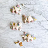 The pastel felt unicorn garland is displayed on a white marble surface, creating a whimsical atmosphere. This unique wool felt garland features four white unicorn figures, each with pink hooves, pastel yellow, blue, and purple manes, and delicate felt wings with pink embroidery. A thin black string adorned with small white felt balls connects the charming unicorns.