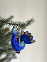 A vibrant felt peacock ornament is nestled among green Christmas tree needles, set against a soft white background. This handcrafted felt ornament features a rich blue body, a yellow beak, and a fanned tail adorned with blue and gold accents. Tufts of green, blue, and purple feathers crown its head, while a jute string is attached for hanging.