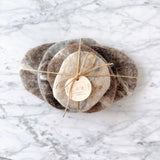 This set of five handcrafted wool felt pebble stone coasters is displayed on a light marble surface. Each unique, irregularly shaped coaster is made from organic wool felt in varying earthy tones, evoking rustic charm. The set is beautifully bundled with natural hemp string and features a simple logo tag, highlighting its fair-trade craftsmanship by Nepalese female artisans.