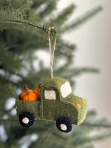 The Felt Pickup Truck with Pumpkins Ornament, a charming handcrafted felt ornament, dangles by a jute string from a green pine tree branch. The green felt truck carries bright orange pumpkin ornaments in its bed. It features white felt windows, yellow felt headlights, and black wheels with white felt hubcaps, evoking a playful autumnal mood.
