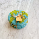 Four handcrafted felt Planet Earth coasters are neatly stacked on a marbled white surface, tied with a jute string and a small tag. Each round, wool felt coaster features a light blue base with textured green continents, creating a miniature world design. These unique, eco-conscious coasters highlight their artisanal quality.