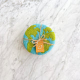 A set of four organic wool felt Planet Earth coasters rests neatly bundled on a white marble surface. Each round, handcrafted felt coaster features a light blue ocean base with green continents, evoking a global design. The ethically made set is tied with a rustic jute string bow and a small 'Ganapati Made in Nepal' tag, highlighting its fair-trade craftsmanship.