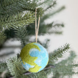 A spherical felt planet ornament hangs from a verdant pine branch, highlighting its eco-conscious design. This needle felted wool ornament features a detailed world map with textured green landmasses and a white gradient tie-dye pattern representing water, adding a unique, handcrafted charm suitable for festive displays.