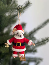 A festive wool felt Santa mouse ornament hangs prominently on a blurred Christmas tree. This handmade felt ornament features a cute mouse dressed in a red and white Santa suit with a black belt and gold buckle. The mouse has beige ears, a white face with black whiskers, and small black eyes, holding a small gold bell in its left hand. A jute string is attached to its hat for easy hanging.