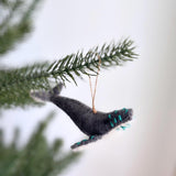 A gray wool felt whale ornament, part of a set of felt sea animal ornaments, hangs on a Christmas tree. This handmade felt whale ornament features a gray body with a turquoise-embroidered tail. A visible jute string on its back allows for hanging, showcasing its detailed texture and craftsmanship.