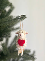 A charming handmade felt sheep ornament, holding a red heart, is displayed on a Christmas tree. This white wool felt sheep features textured wool, pink inner ears, and a stitched smile. A jute string is attached for easy hanging, adding a touch of warmth and affection to any decor.