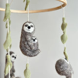Felt Sloth Hugging Baby ornaments are displayed hanging from a wooden mobile frame. The prominent gray and cream felt sloth ornament features a serene expression with closed eyes and a gentle smile, tenderly cradling a smaller baby sloth, also with closed eyes. White cotton strings run through light green felt leaf embellishments, attaching the ornament to the frame. Two additional felt sloth ornaments are partially visible in the soft, light background.