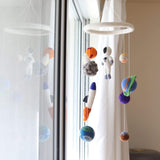 A colorful wool felt spaceman baby mobile hangs suspended in front of a bright window with white curtains, creating a soft, dreamy atmosphere. The mobile features a white felt ring from which a white spaceman, colorful planets, an orange and blue rocket, and a gray asteroid are suspended. Each needle-felted figure, crafted from 100% New Zealand wool, is detailed and textured, offering a whimsical, three-dimensional look.