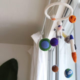 A felt spaceman baby mobile hangs in front of a bright window with sheer white curtains, creating a soft, dreamy atmosphere. The mobile features a circular white felt frame from which 3D needle-felted figures are suspended by clear strings. These wool felt elements include colorful planets in orange, brown, green, blue, and purple, a white spaceman with an American flag, and a white and orange rocket. Its soft textures evoke gentle creativity and comfort for a nursery.