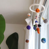 A Felt Spaceman Baby Mobile hangs gently in a bright, airy room, illuminated by natural light from a window with sheer white curtains. This wool felt baby mobile features colorful, 3D felted figures of orange, white, purple, and blue planets, a rocket, and an astronaut, suspended by clear strings. A large green houseplant adds a touch of nature on the left.