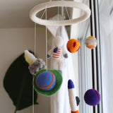 A whimsical felt spaceman baby mobile is displayed in a brightly lit room. It features a white felt ring from which various needle-felted space-themed figures hang by thin white strings. These include an astronaut with an American flag, an orange and blue rocket, and vibrant spherical planets, like one orange with red spots and another blue with an orange ring. The playful design evokes a sense of wonder.