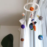 A colorful wool felt spaceman baby mobile hangs suspended in front of a bright window. This unique baby mobile features needle-felted 3D figures, including a white astronaut with an American flag, various planets in orange, purple, brown, and blue, and a green and blue rocket, all suspended from a white felt ring.