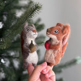 Two handcrafted needle felted squirrel finger puppets, known for their eco-friendly design, are playfully held by a female hand against a blurred green pine tree. These unique felt squirrel finger puppets, made by artisans, feature one gray squirrel holding a tiny brown acorn and another reddish-brown squirrel clutching a bright red nut. Both showcase small black eyes and intricate textures, perfect for imaginative woodland storytelling.