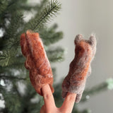 Two handmade felt squirrel finger puppets are worn on a hand, held against a blurred Christmas tree background. Crafted from soft, natural wool by artisans, one warm brown squirrel holds a tiny felt acorn, and the other, a cooler gray-brown, carries a bright red nut. These unique and eco-friendly felted finger puppets are perfect for imaginative woodland storytelling.