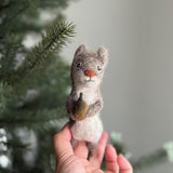 A handmade, needle-felted wool felt squirrel finger puppet, holding a tiny brown felt acorn, is held in a female hand. This charming squirrel puppet features a textured, grayish-brown body with a contrasting white belly. The soft, neutral backdrop includes a blurred Christmas tree, highlighting this unique handcrafted felt kids toy.