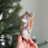 A handmade felt squirrel finger puppet, crafted from wool felt, is held in a female hand against a softly blurred Christmas pine tree background. The gray squirrel puppet features a reddish-brown bushy tail and clutches a small brown acorn in its paws. This charming, needle-felted toy highlights intricate artisan detail, offering an eco-friendly option for imaginative play.