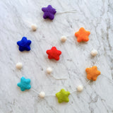 A cheerful wool felt star garland rests on a marble surface. This vibrant garland features seven soft, tactile felt stars in a rainbow of purple, blue, teal, green, red, orange, and yellow. White felt balls are evenly spaced between each star, all connected by a thin white string.