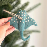 A blue wool felt stingray ornament with hemp string, handcrafted by Nepalese artisans, hangs on a Christmas tree. This fair-trade, needle-felted piece features white polka dots, two small black eyes, and a curled tail, adding festive charm.
