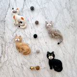 A cheerful wool felt garland featuring stitched cats rests on a marbled white surface. This handmade garland displays four 3D-designed cats in black, orange, grey, and white. Small, alternating colored felt balls string the cats together, with a copper bell attached by a tiny felt ball at one end.