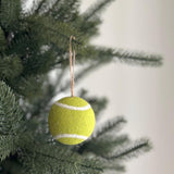 A wool felt tennis ball ornament hangs gracefully from a green Christmas tree branch. This handcrafted sports Christmas decoration is bright yellow with a white line encircling its center, accurately mimicking a real tennis ball. A natural jute string is attached, perfect for adding a playful touch to your holiday decor.