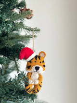 A whimsical felt tiger Christmas ornament, wearing a Santa hat, hangs on a green Christmas tree with a soft white background, creating a festive atmosphere. The felt tiger ornament has orange and black stripes, a fluffy white belly and muzzle, and a classic red and white Santa hat atop its head. A jute string is visible for hanging.