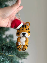 A handcrafted wool felt tiger ornament with a red Christmas hat is delicately held by a female hand near a festive Christmas tree. The charming tiger features orange and black stripes, a cream belly and face, tiny black eyes, and a sweet smile, embodying a whimsical and celebratory atmosphere.