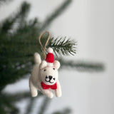 A charming handmade felt white cat Christmas ornament, wearing a festive red Christmas hat and a matching red bow tie, hangs from a pine branch by a jute string. The delightful felt cat ornament is presented against a blurred background of a Christmas tree and a plain white wall, ready for holiday display.