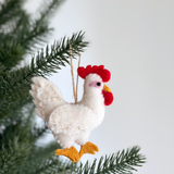 A plump, white felt chicken ornament hangs from the green needles of a Christmas tree. This handmade wool felt chicken features a bright red crest, a yellow beak, and orange feet, all intricately needle felted. A jute string is visible for hanging.