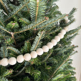 A White Pompom Ball felt garland is draped across the green branches of a Christmas tree in a close-up shot. The festive felt garland features a series of evenly spaced, round, off-white felt pompom balls, creating a cozy winter holiday ambiance.