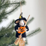 A whimsical wool felt wizard mouse ornament is showcased against a blurred Christmas tree backdrop. This handcrafted white mouse is dressed as a witch, wearing a black cloak and a pointy black hat detailed with a yellow crescent moon. It charmingly holds a small, smiling orange pumpkin and a miniature witch's broom.