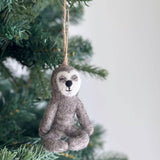 A gray felt yoga sloth ornament, suspended by a jute string, hangs from a Christmas tree branch. The unique Christmas ornament features a white face, a small black nose, and a small black mouth. It is depicted in a meditative seated pose with its eyes gently closed, creating a peaceful and festive mood.