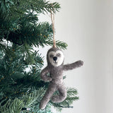 A felt yoga sloth ornament hangs from a Christmas tree, evoking a peaceful, festive mood. This grey felt sloth ornament features flexible wire in its limbs, allowing it to be posed in various yoga positions. It is detailed with a white face, a black nose, and a jute string for hanging.