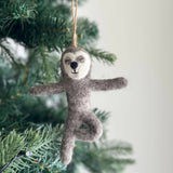 A handmade felt Yoga Sloth ornament hangs serenely from a lush green Christmas tree branch. This charming gray felt sloth, with a white face, black nose, and closed eyes, features flexible wire limbs posed whimsically with outstretched arms and legs. A jute string is attached to its head, allowing it to gracefully adorn the tree, adding a peaceful and festive touch.