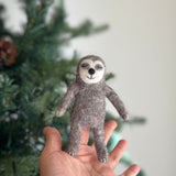 A handmade felt sleepy sloth finger puppet is held gently in a female hand, set against a softly blurred Christmas tree. This gray and light-faced sloth puppet features closed eyes, a gentle smile, and tiny white-stitched claws on its arms and legs, highlighting its tactile charm.