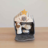A handmade felt finger puppet set, featuring a nativity scene with 12 intricately crafted figures, including Baby Jesus, Mary, Joseph, three kings, an angel, a shepherd, a cow, a sheep, and a donkey. The set is presented in a felt stable with an open door, placed on a wooden table with a white surface, evoking comfort and tradition.