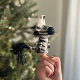 A handcrafted needle-felted zebra finger puppet is playfully presented, held by a female hand with dark red nails against the soft, festive glow of a Christmas tree. This unique felt puppet features iconic black and white stripes, a tufted mane, and a black tail, embodying a cozy holiday spirit.