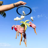A hand holds a wool felt baby mobile featuring three flamingo figures hanging from a floral design, set against a blurred outdoor background under a blue sky. The product showcases a whimsical and playful design with a tropical theme.